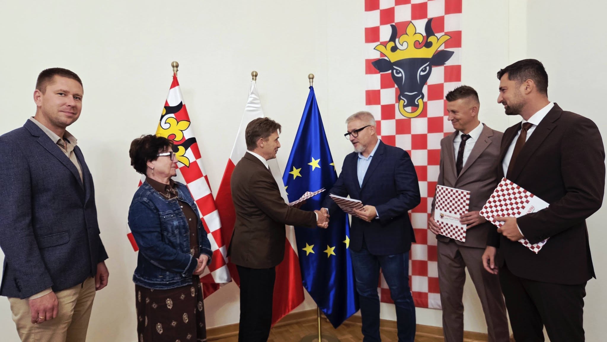 przedstawiecie władz samorządowych z firma która będzie wykonywąła budowe sciezki rowerowej, za tymi osobami widoczna ściana i flaga powiatu kaliskiego